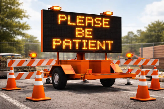 Orange led message board on a trailer displaying ’please be patient’ in glowing yellow letters, flanked by orange and white striped traffic barriers and cones.