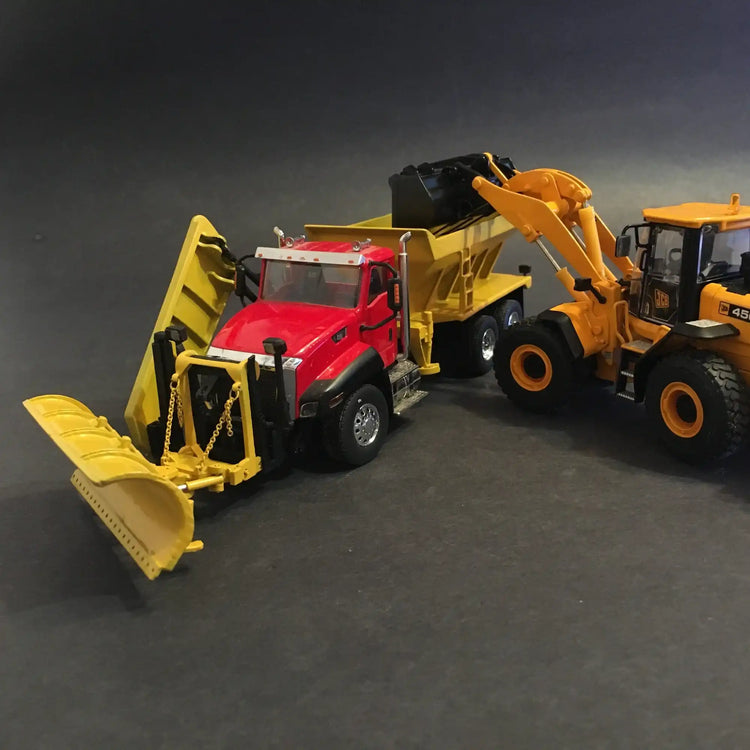 Red dump truck with yellow front-mounted plow attachment and yellow construction equipment nearby.